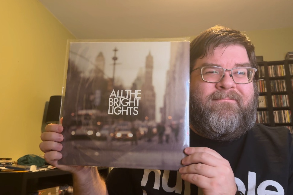 Photo of a white man holding with brown hair and brownish-gray beard the All The Bright Lights album in a plastic sleeve up next to my head and smiling. I'm wearing a black shirt with white writing and there's a bookshelf behind me on my right and a lower shelf with a lamp and some random junk on it.