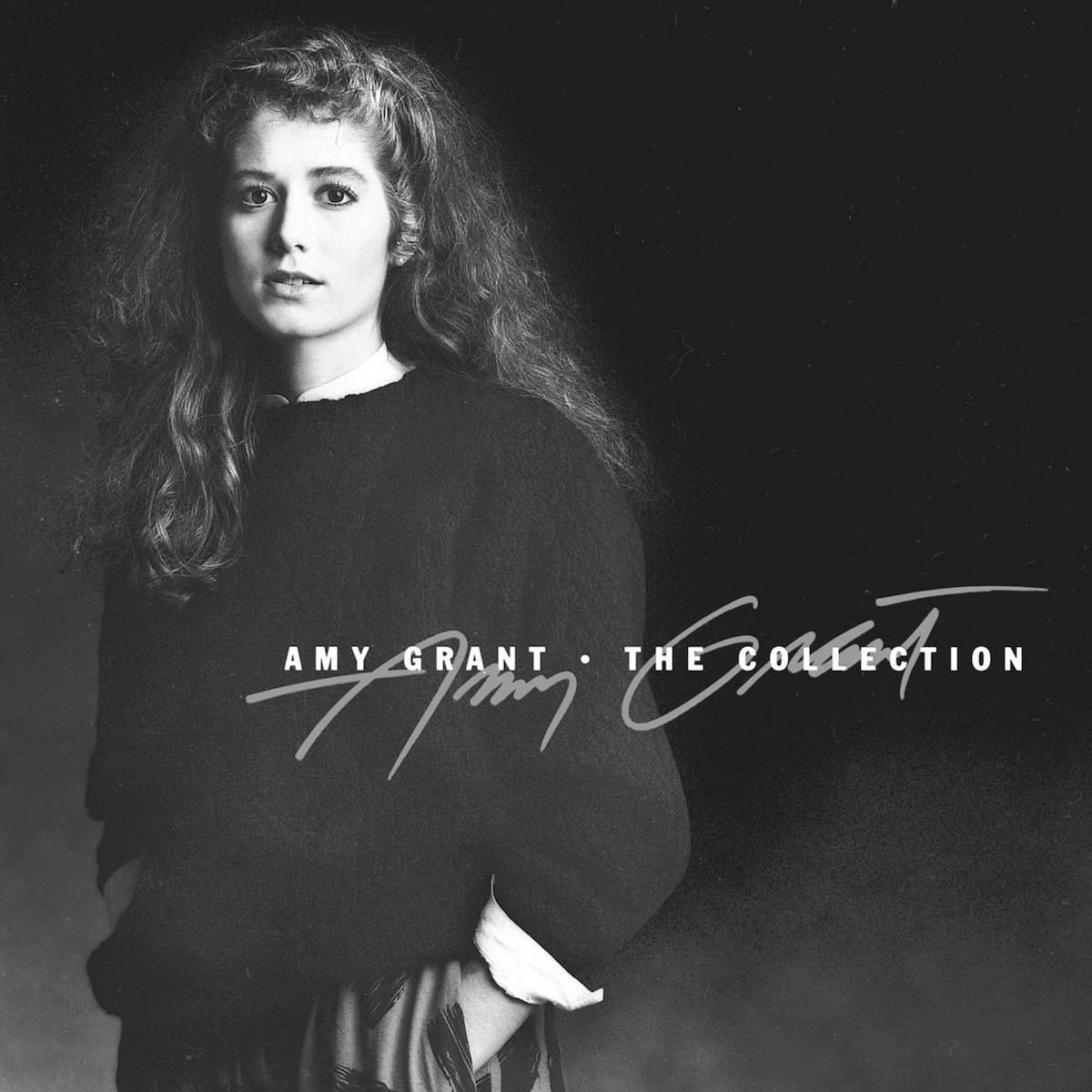 Album Art of Amy Grant's The Collection album - On a black background with a bit of a grey light at the bottom, a black-and-white photo of a young, white woman with long, curly hair is looking at the camera with a rather blank look. She looks to be wearing a dark sweater over a white shirt with a lighter skirt or pants below it. She's photographed from the waist up and has her hand in a pocket. Printed on top of the photo in grey is the signature of the artist, with in white text in a non-serif font above it is the artist name, a dot, and then the album title.