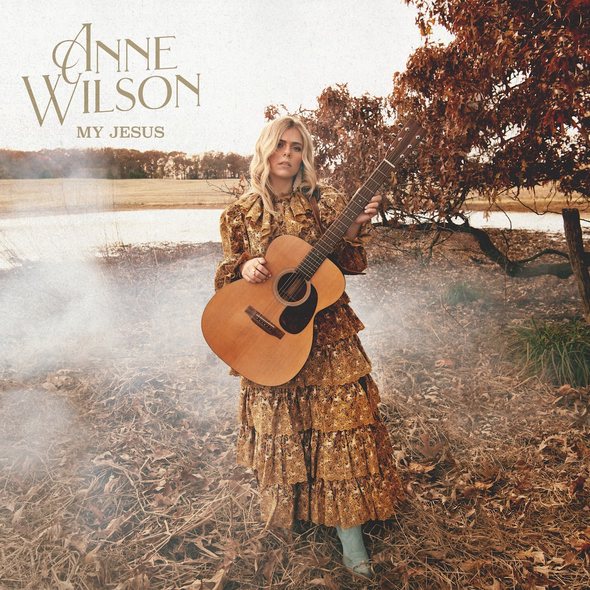 Album Art of Anne Wilson's My Jesus album - A color photo of a young, white woman with shoulder-length blond hair standing in some brown grass and leaves with a pond behind her and a tree with brown leaves to the right, and off in the distance is a brown grass field and a line of trees before the horizon. The sky is a bright grey, not blue, so seems to be a thick layer of clouds. The woman is standing in the foreground with a golden brown, frilly dress on and holding an acoustic guitar. In the top left, printed above the sky is the artist's name in large gold letters with fancy capitals on each first letter and then much smaller below is the album title.