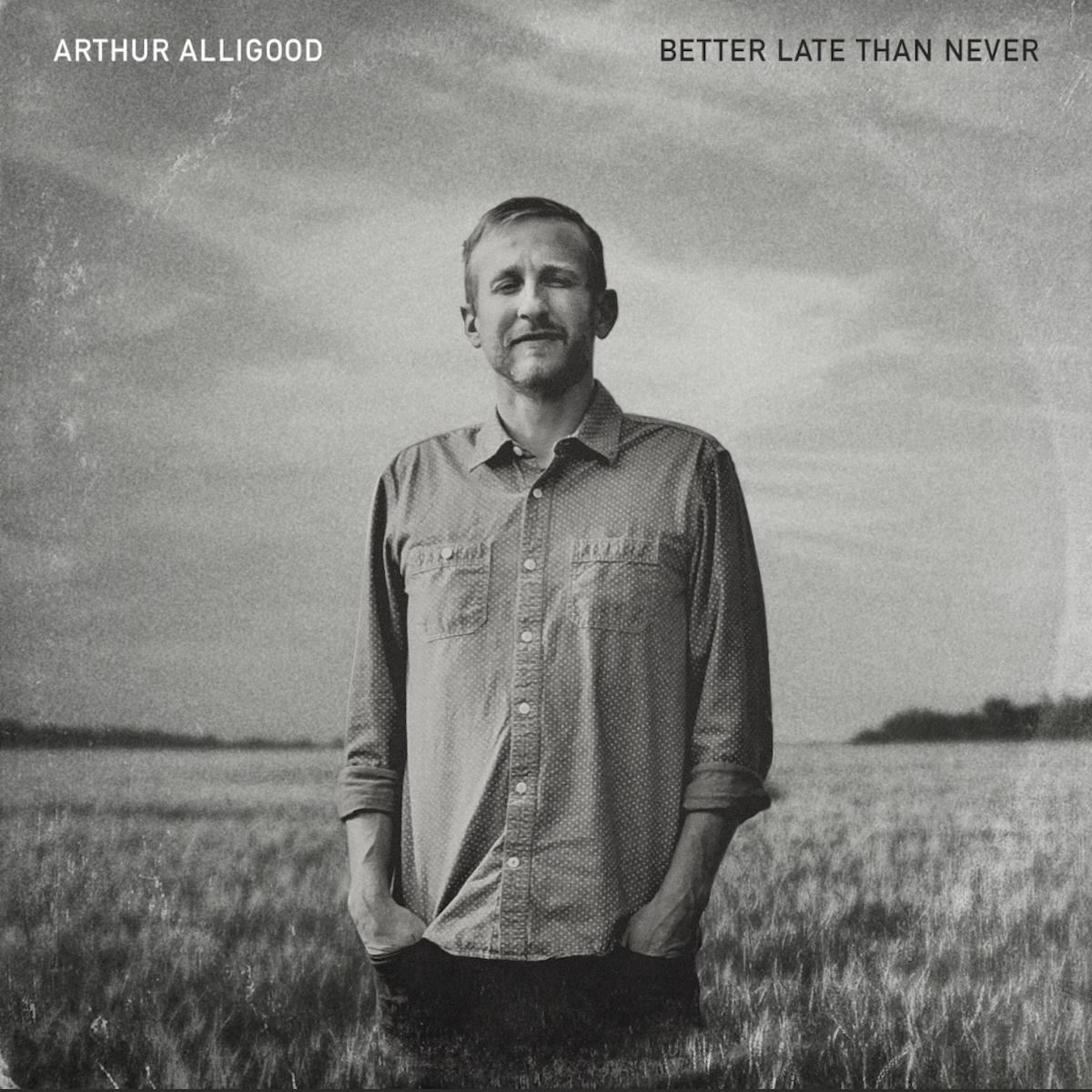 Album Art of Arthur Alligood's Better Late Than Never album - A black-and-white photo of a middle-aged white man standing with his hands in his pants pockets in a field of long grass. The field and sky are a gray color, and on the horizon some trees that are black. The man is pictured from the legs up, with dark pants and a light-colored collared shirt with pockets on both sides. He's god short hair and a short beard and mustache of a light color. The picture has simulated ringwear with a circle where the vinyl would be inside a package. In the top left, printed in white is the artist name, and in the top right, printed in black is the album title.