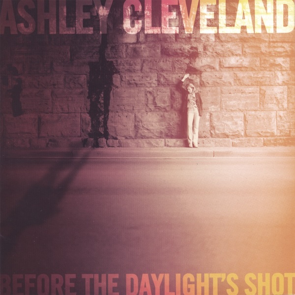 Album Art of Ashley Cleveland's Before The Daylight's Shot album - A photo of a road in the foreground and a large, brick retaining wall just past the road. Standing in front of the wall is a white woman with long, light-colored hair, holding up her right hand above her head and looking down to the roadway at her feet. The right side of the image is overexposed, like maybe there was too much sunlight over there, but the left side is very dark. Printed in transparent white text right across the top edge is the artist's name in large text, and along the bottom is the album title in smaller text.