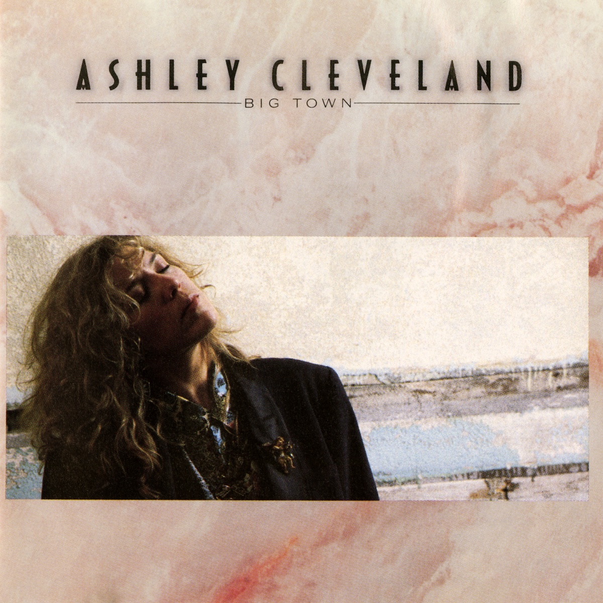 Album Art of Ashley Cleveland's Big Town album - The background looks like some sort of pink and white marble stone. Across the bottom of the middle is a wide photo of a young woman with long, light brown curly hair. he's wearing a black coat and and has her eyes closed and has a contemplative look on her face. She seems to be sitting next to a wall with some odd horizontal holes in it. At the top, on top of the marble it has the artist's name printed in large, black letters, and then a hairline with the album title in much smaller letters in the center of the line.