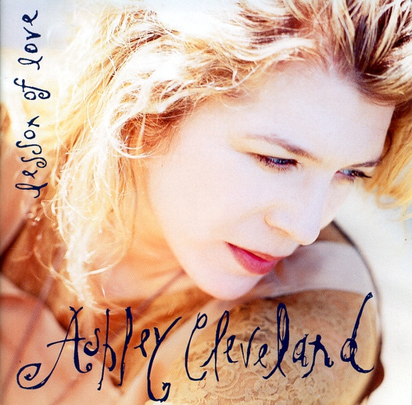 Album Art of Ashley Cleveland's Lesson Of Love album - A photo of a young, white woman's face. She has blonde-brown hair almost down to her shoulders and is photographed from above, it seems. Below her face, her name is written in dark blue, very handwritten-looking writing. On the left side vertically, the album title is also written in smaller but also handwritten text.