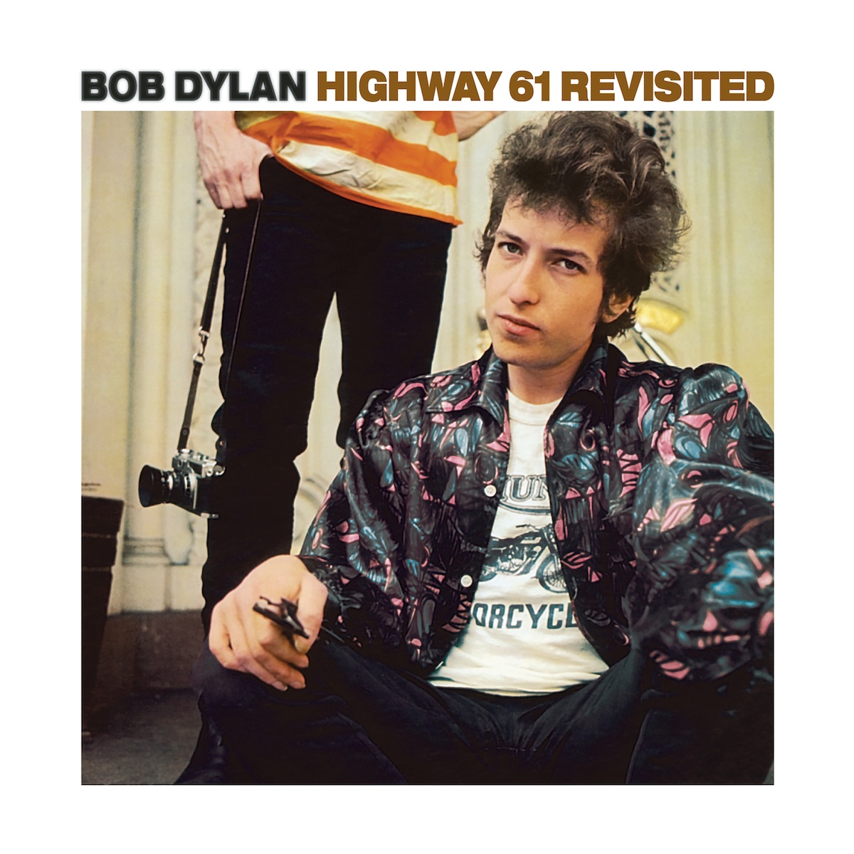 Album Art of Bob Dylan's Highway 61 Revisited album - With a generous white border around it, a color photo of a young white man with curly hair at least a few inches long is looking somewhat annoyedly at the camera. He seems to be sitting down because someone's legs and waist can be seen behind them with black pants and an orange-and-white striped shirt and holding a camera on a strap. The young man pictured is wearing a black, blue and pink colored shirt with a fun pattern and it's open in the front to reveal a white T-shirt underneath with a motorcyle on it. Above the photo, in the white border area, it says the artists name in black followed by the album name in brown letters. The lettering is all caps.