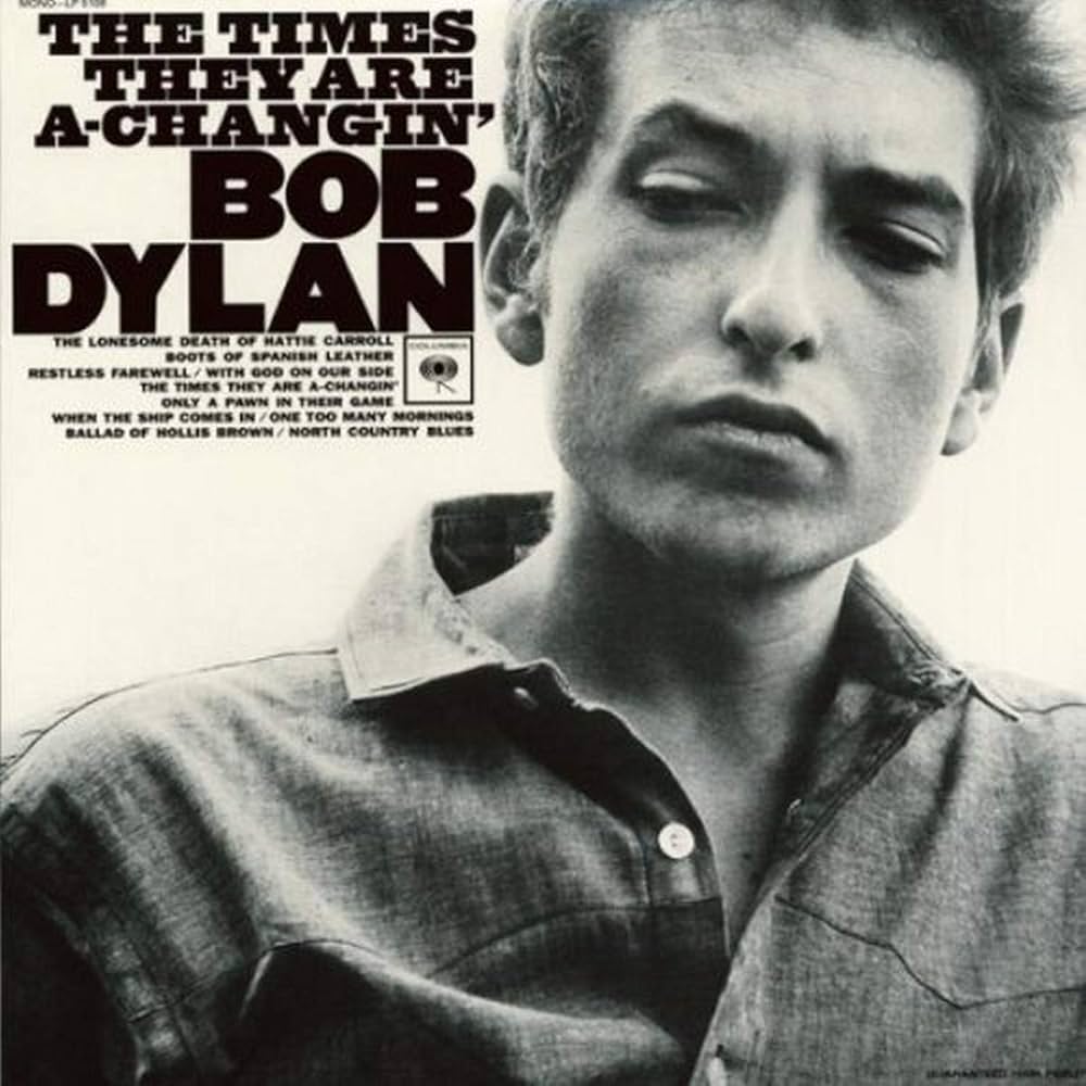 Album Art of Bob Dylan's The Times They Are A-Changin' album - A black and white photo of a white man from shoulders up wearing a collared shirt and looking down and to the left in a bit of a sour, skeptical look. Next to his face, the album title and artist name in large black letters, with the Columbia Records logo and track listing in smaller black print below.