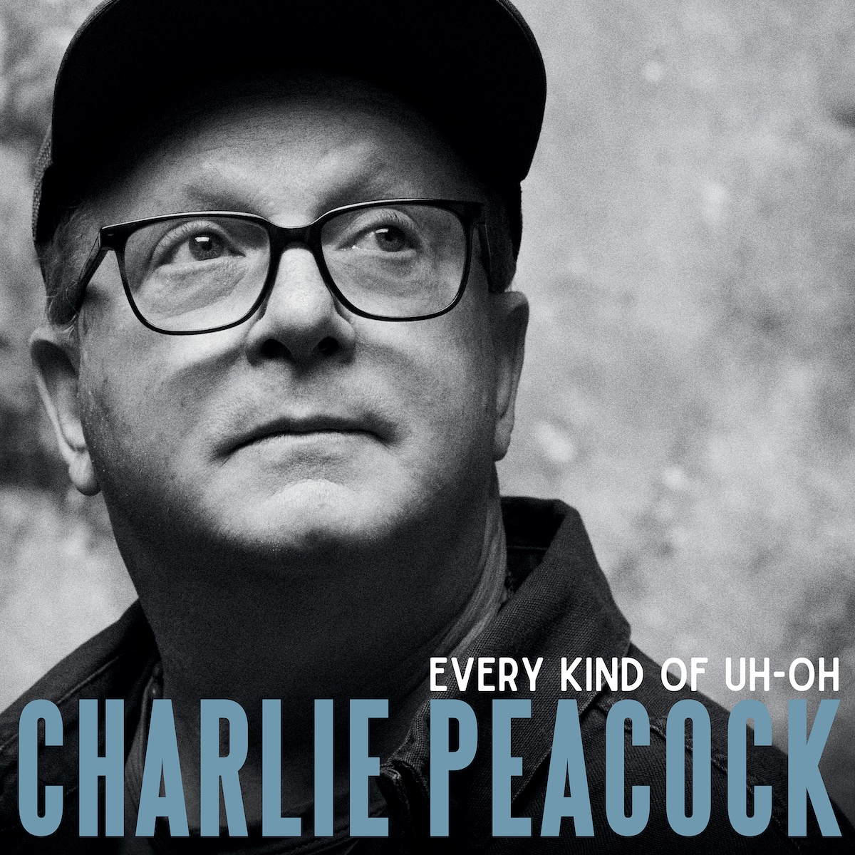 Album Art of Charlie Peacock's Every Kind Of Uh-Oh album - A black-and-white photo of a white man wearing dark-rimmed glasses and looking up and to his left. He's pictured from just the shoulders up, is wearing a baseball cap which is shading his forehead, and the background is a blurry gray image. Printed on the bottom in large light blue letters it he artist's name, and above it to the right in smaller, white letters is the album title.