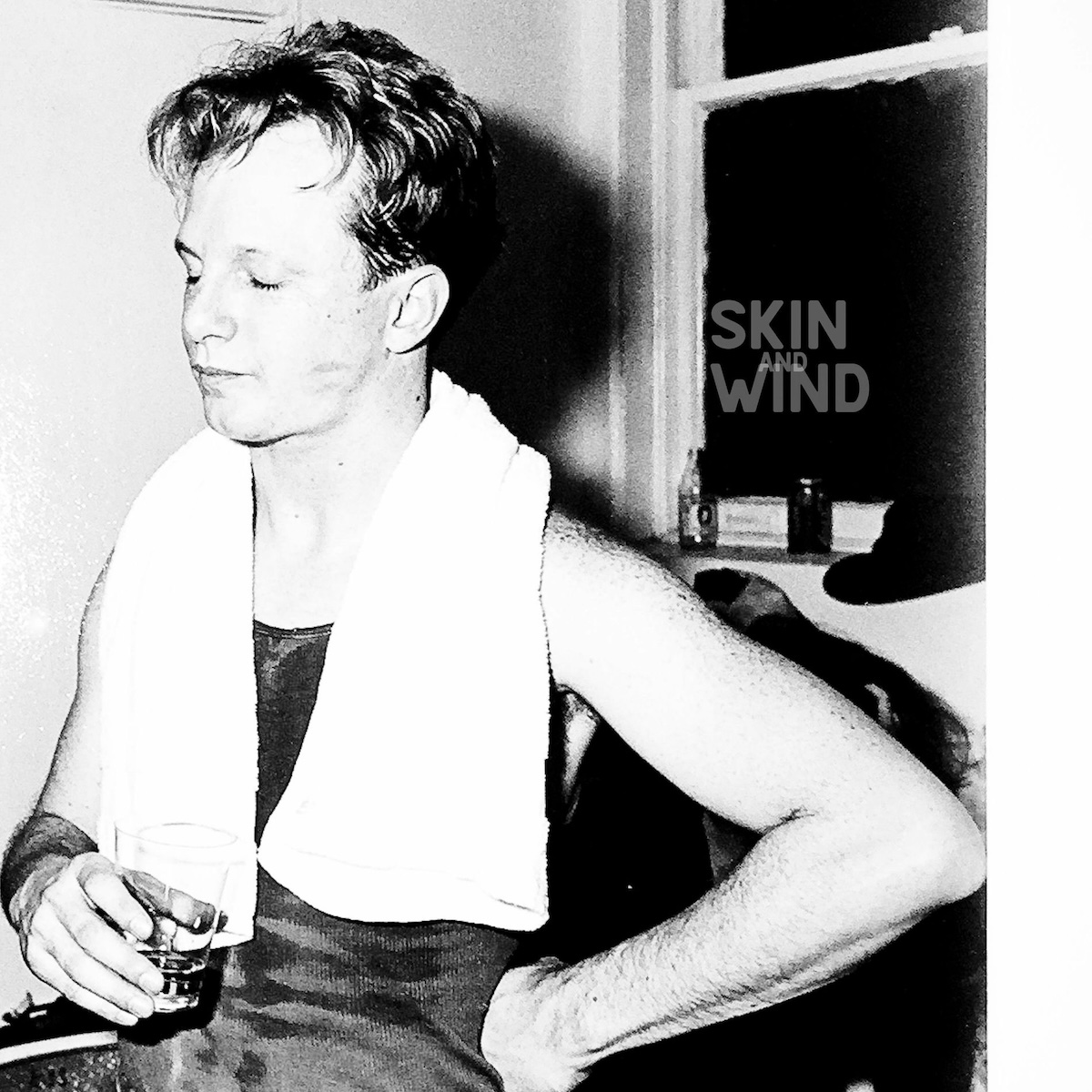 Album Art of Charlie Peacock's Skin And Wind album - A black-and-white photo of a young man standing with his eyes closed and a glass in his hand. His other hand is on his hip and his arms are bare, and it looks like he's been boxing or working out because he has a white towel around his neck. Behind him can be seen a window with a hat and a bottle and can of different drinks. On the window, printed above the photo is the album title, with 'and' being very small and overlapping the word 'wind'.