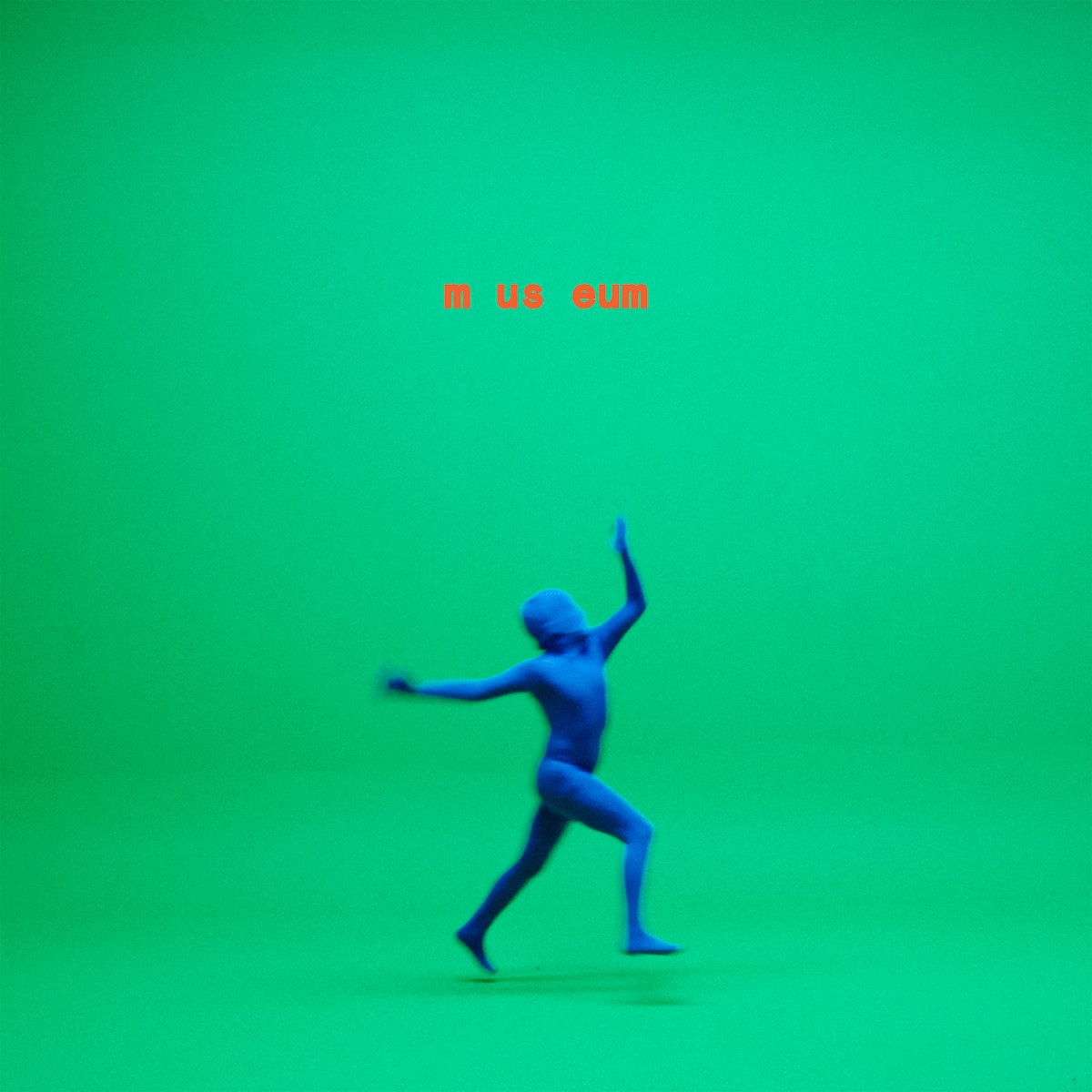 Album Art of Citizens's Museum album - A photo of a large green screen room with both green walls and a green floor and no real easy-to-spot place wher the wall and ends and floor starts. On the floor is a person covered from head to toe in some sort of blue skin-tight suit. They are caught in motion in the middle of the photo, it looks like dancing or something, and both of their feet are off the floor because there's a shadow below them. Above the blue person's head, printed in orange is 'm us eum' in a bold, orange font.