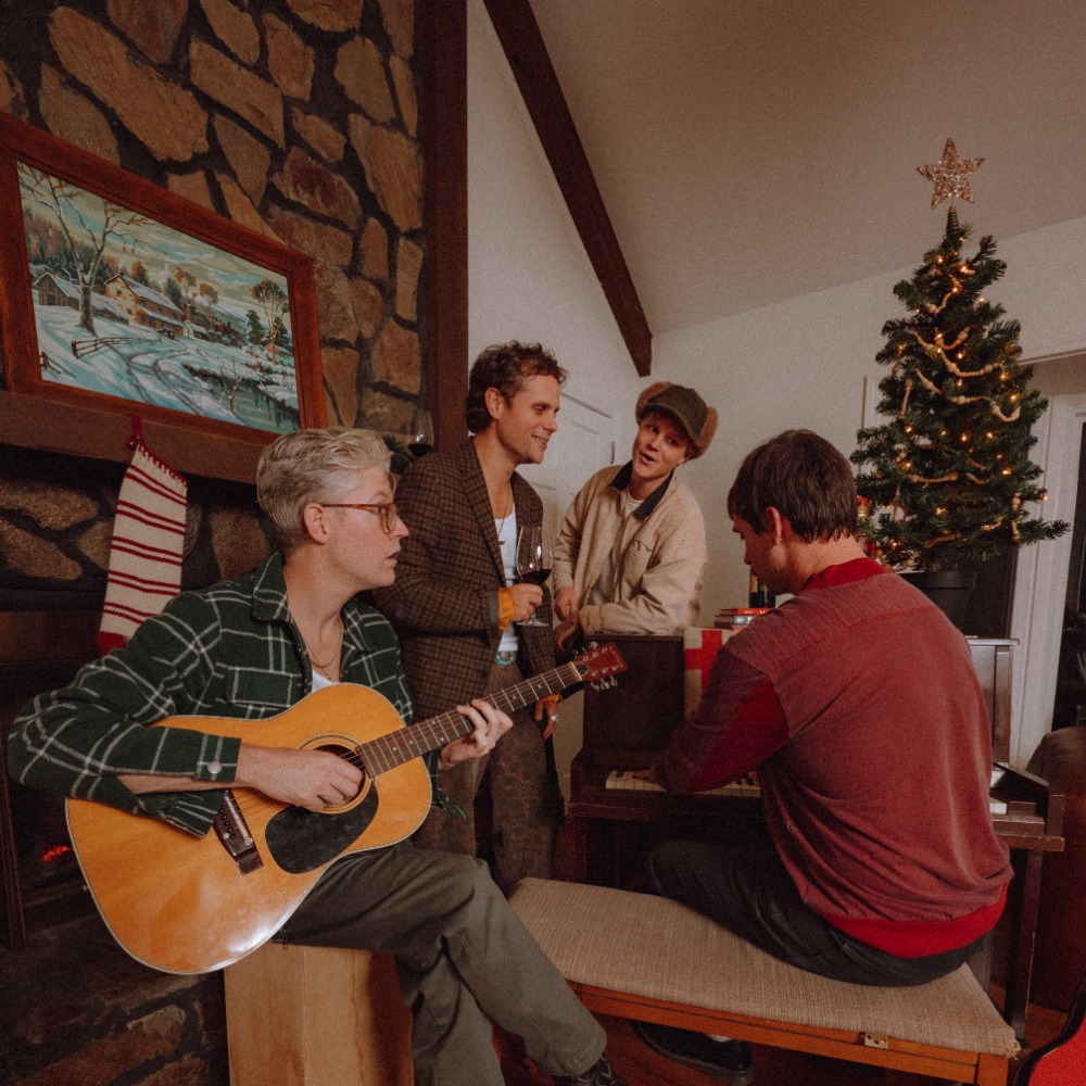 Album Art of Colony House's Every Christmas EP - Four white men are gathered in front of a fireplace with stones stretching up to the ceiling. Two are standing in the back, while one is playing an acoustic guitar and sitting on the box in front and the fourth is not facing toward the camera, but instead facing away and playing a piano. There is a small Christmas tree behind the piano and some wrapped presents on the piano.