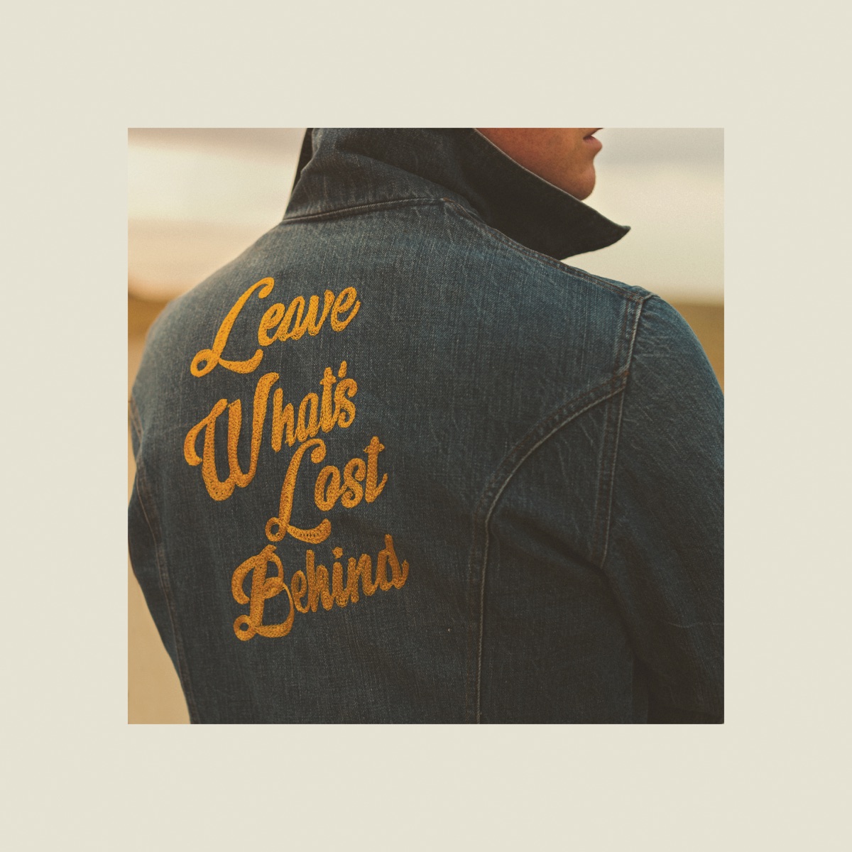 Album Art of Colony House's Leave What's Lost Behind album - surrounded by a wide, beige border is a photo of the back of a person. We can only see the bottom of his face, but the photo focuses on a denim jacket which has the words 'Leave What's Lost Behind' stitched in gold on the back. It looks like the person may be outside on a cloudy day about golden hour.