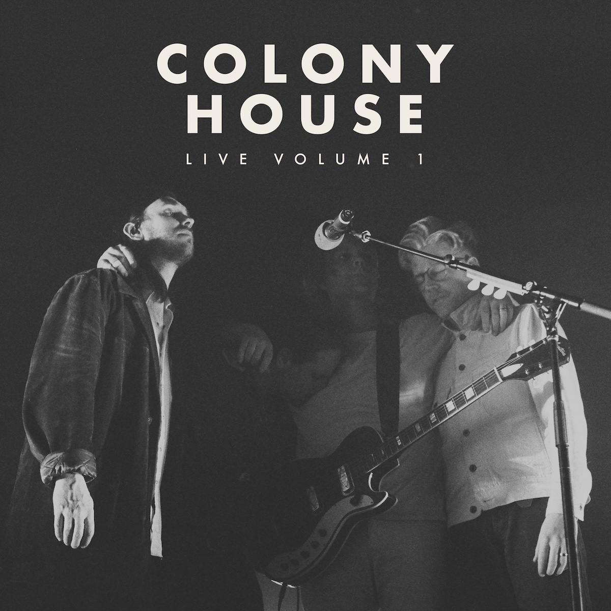 Album Art of Colony House's Live Vol. 1 album - A black-and-white photo of four men huddled together around one microphone in a mostly dark area, definitely very black behind them and mostly lit from the right side. The two men on the outsides are the most visible, and they are both wearing button-down shirts and dark pants. The person close to the microphone is mostly in the dark, but has a guitar hanging around his neck and he's not playing it, he's got his arms around his bandmates. In the middle mostly in the dark is one of the young man with his face in the chest of his band-mate, his brother. In the darkness above them is printed the band name in large, bold letters in a creamy white. Below it much smaller letters it says 'Volume 1'.