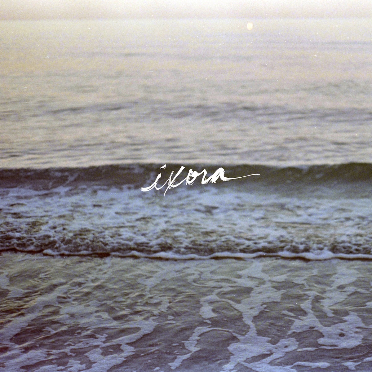 Album Art of Copeland's Ixora album - A color photo of a wave about to break on a beach. The wave is hitting its peak at the middle of the picture vertically, and the froth of the waves before it is below it on the sand. Behind the current wave is out of focus, but you can see small waves behind it as well and the horizon and light of the sun out of frame above. In the middle, printed on top of the photo, is the word 'ixora' painted in white paint with a flowing hand.