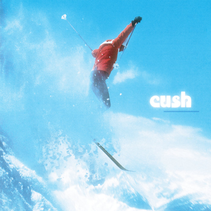 Album Art of Cush's self-titled album - In front of a bright, blue sky, a man in a red jacket is skiing down a hillside and is photographed from the side. The person is looking down and is flying through the air, kicking up snow like he probably just jumped off some part of the hill. His hands and poles are in the air, and below his skis you can see far-off a beautiful mountainside and even clouds. Printed on top of the blue sky on the right side is the word 'cush' in white, with a darker blue line below it offset slightly to the right.