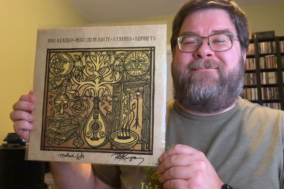 A photo of a rotund white man from the shoulders up with a light-colored beard and mustache, darker brown hair, and large glasses smiling at the camera and holding up a record jacket of the Strings & Sonnets album.