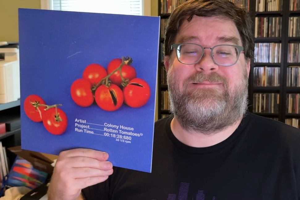 A photo of a middle-aged white man's face smiling slightly painfully into the camera, and next to his face, he's holding the Colony House Rotten Tomatoes 12-inch vinyl in its blue and red paper sleeve. He's got a short beard, is wearing a black shirt and has shelves of CDs behind him.