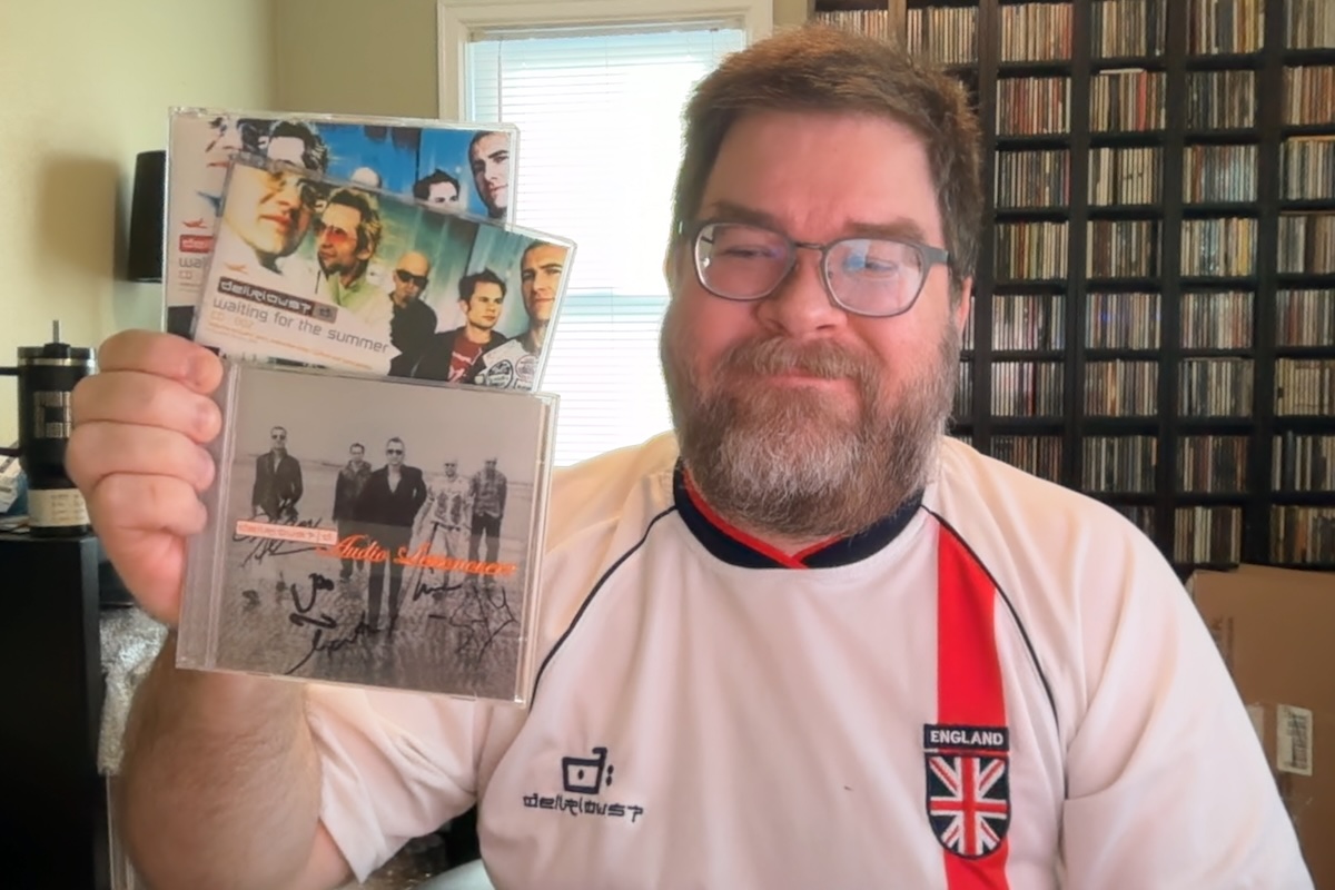 A photo of a middle-aged man with short hair and a short beard in a room with large CD racks and other junk behind him. He's smiling and wearing a Delirious? soccer jersey and holding 3 CDs: the Audio Lessonover? CD with a signed booklet and two 'Waiting For The Summer' singles.