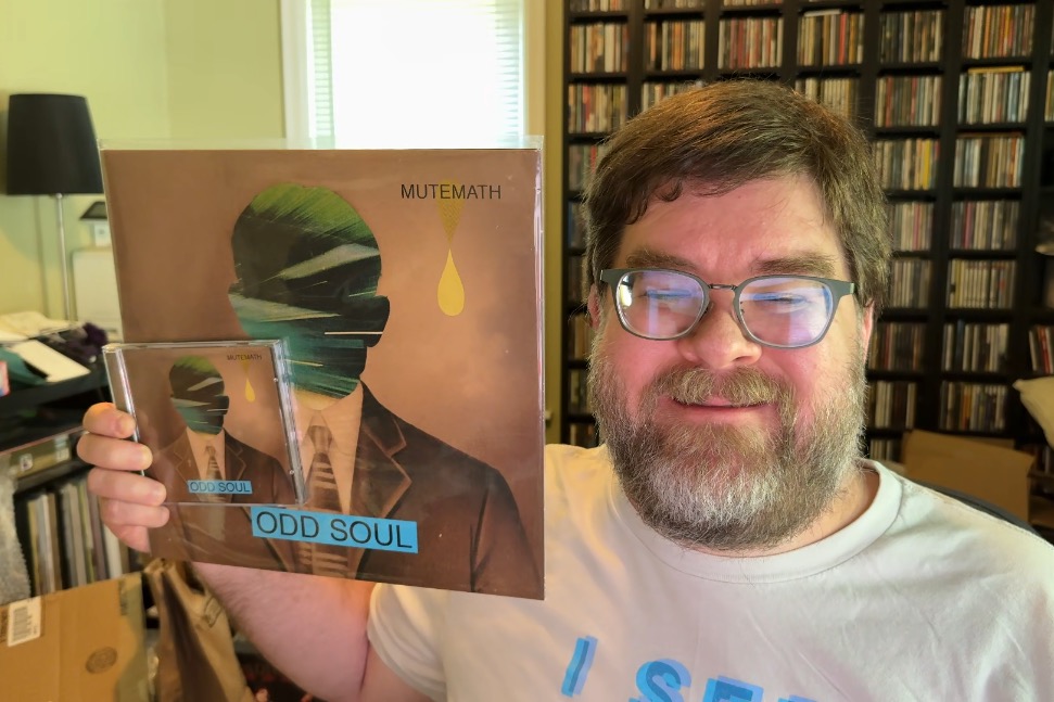 A middle-aged, rotund man is shown from the shoulders up holding a vinyl record and CD of this album in his right hand. He's got short hair and a full beard that's also pretty short, and glasses that are showing some refection from the monitor. He's smiling and has a rack of CDs and some vinyl and some boxes behind him.