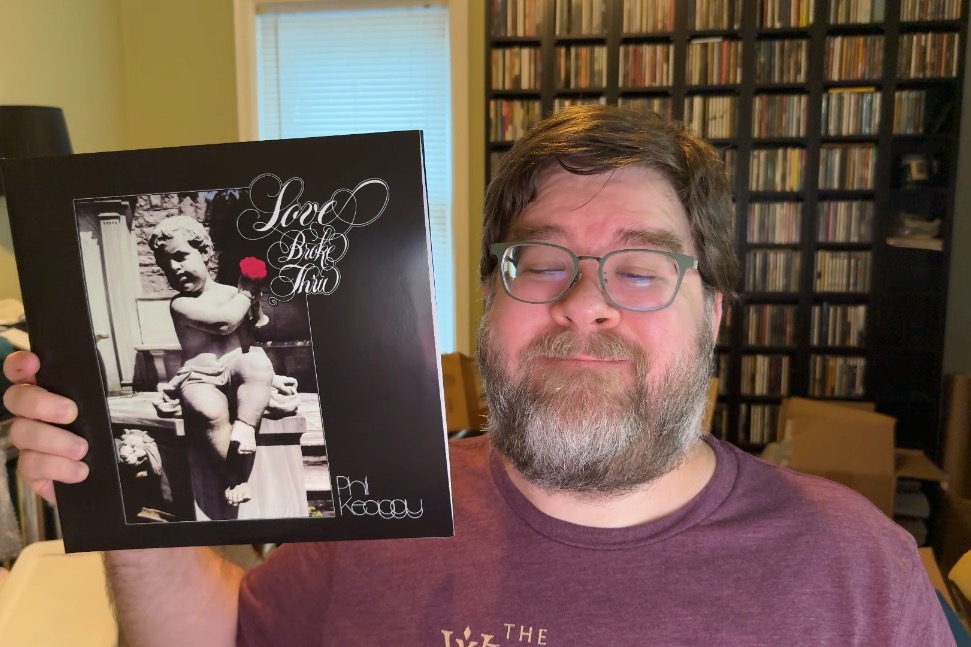 From the shoulders up, a middle-aged rotund man wearing a dark red T-shirt is holding up a vinyl record in a cardboard slipcase that looks just like the Love Broke Thru album cover. Behind him is a room with a warm vibe, CD racks off to the right, and lot of random messiness. The man has glasses, a beard and a mustache and is smiling.