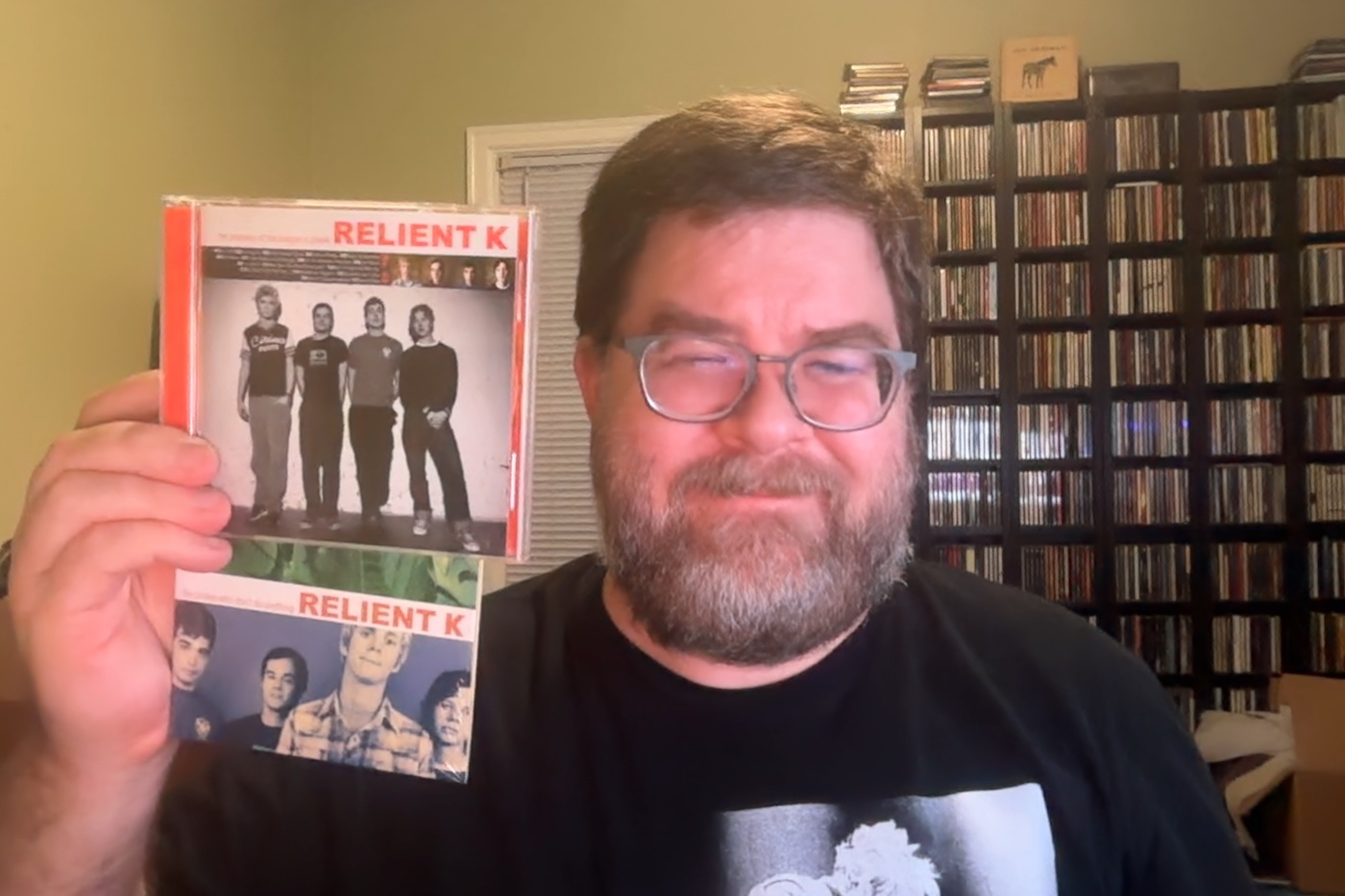 A photo of a rotund, white man from the shoulders up who has a full beard and mustache. He's wearing a black shirt and smiling and has in his right hand two CDs from Relient K, The Anatomy Of The Tongue In Cheek album and the Pirates Who Don't Do Anything single. Behind him is a large wall of shelves with CDs on them and a window frame.