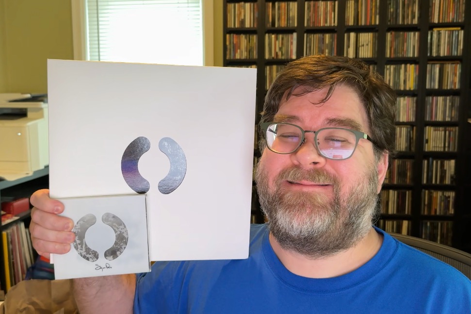 A photo of a rotund white man from the shoulders up, wearing a bright blue T-shirt and having a beard and mustache. He's holding a CD in a jewel case with a parenthesis cut out of the plastic out sleeve. He also is holding in the same hand a larger LP-sized white box with a similarly cut parenthesis hole showing some black-and-white art inside the cutout area.