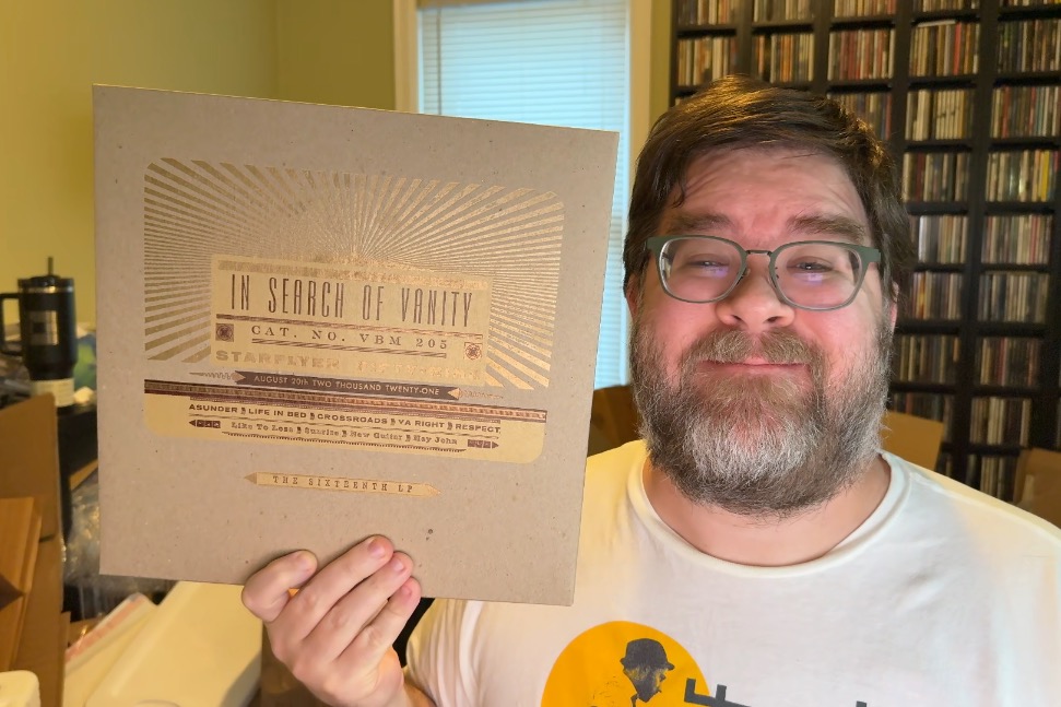 From the shoulders up, a middle-aged rotund man wearing a white T-shirt is holding up a vinyl record in a paper slipcase that looks just like the album cover, but has gold lettering and golden rays that are shining in the light. Behind him is a room with a warm vibe, CD racks off to the right, and lot of random messiness. The man has glasses, a beard and a mustache and is smiling.
