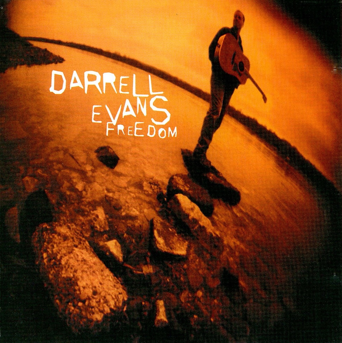 Album Art of Darrell Evans's Freedom album - A very wide-angle lens shows a black-and-white photo where the white is replaced with a gold color. The majority of the photo is of some water and a series of large stones jutting out into the water. A shoreline can be seen on the edges. On the farthest stone out stands a man holding an acoustic guitar, and due to the wide angle, he's somewhat distorted as he's to the top and edge of the frame. In a very jagged-edged font, the artist's name is in white on the left, with the album title just below it a bit smaller.
