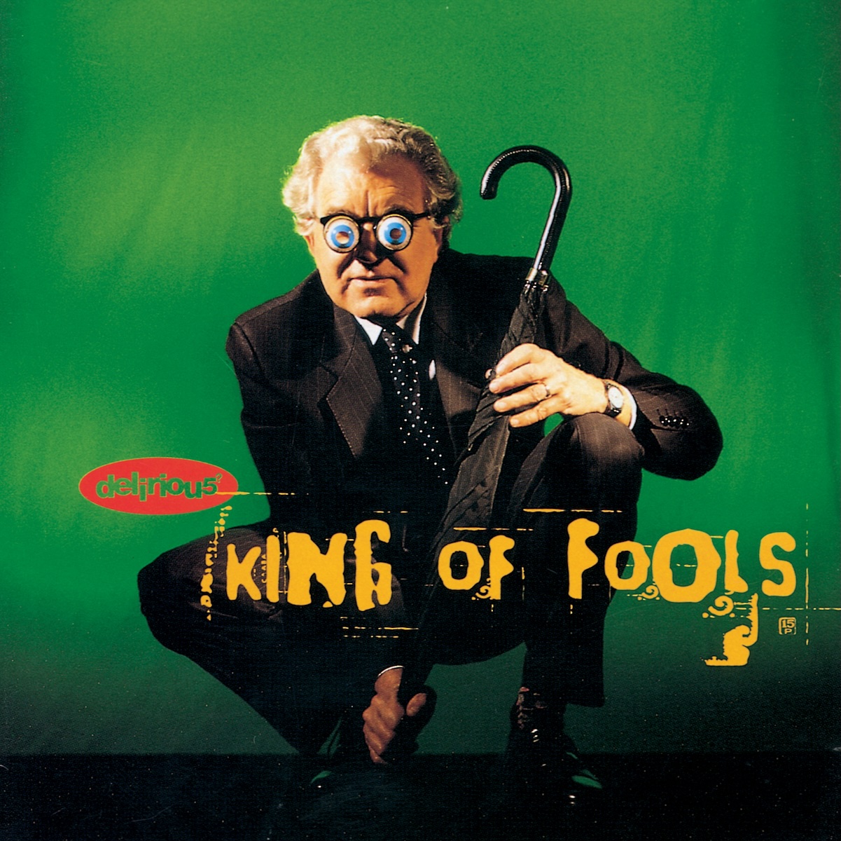 Album Art of delirious?'s King Of Fools album - In front of a green background, an older man with grey hair is crouching wearing a black suit and trousers. He's holding a black, closed umbrella between his legs, kinda in a defensive position. He's also wearing glasses with goofy-looking googly-eyes on them. Printed over his crouched legs is the words 'King Of Fools' in a bright orange, with some extra orange around the edges which makes it look like it could be some sort of graffiti stencil. To the left of that, it says 'delirious?' in letters cut out of a red oval. The '?' is very small and the 's' is a number 5.