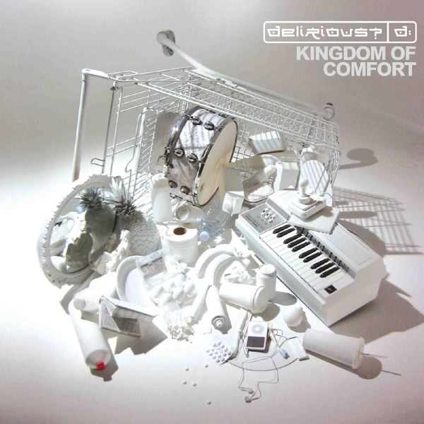 Album Art of delirious?'s Kingdom Of Comfort album - On a gray background, a photo of a white shopping cart that has been tipped on its side towards the camera and is lying on the ground. Spilling out of it is a whole bunch of random things, all colored white. Still in the cart is a snare drum, cartons of milk or other drink, and other foodstuffs. On the right, mostly out of the cart is a small electronic keyboard that is also mostly white. In front of it are soda and coffee cups, an iPod and earphones, pills, plus paper plates, toilet paper, and much more. A mirror is on the left side and has a white painted pineapple, a book or two, and some sort of tube of product with a small cap. In the top right, it has in white the band's logo with the 'd:' word mark. And below that in a light gray is the album cover.