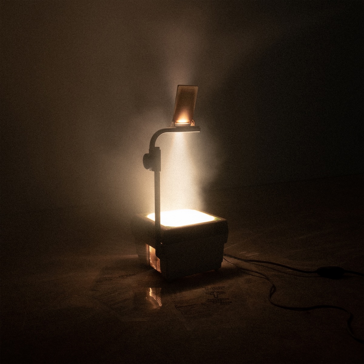 Album Art of Elevation Worship & Maverick City Music's Old Church Basement album - Sitting on a floor in a darkly lit, empty room, the only source of light is an overhead projector box that has s light inside and spills out light from the top, projecting whatever is on the clear plastic sheets placed on top of it onto a wall via a mirror hanging above it. This mostly obsolete technology used to be used in schools for lessons and at churches for communal music before projectors and larger screens became economical. The light from the project in this photo doesn't really illuminate anything visible; it seems the room is a bit smoky, probably for a cool image effect.