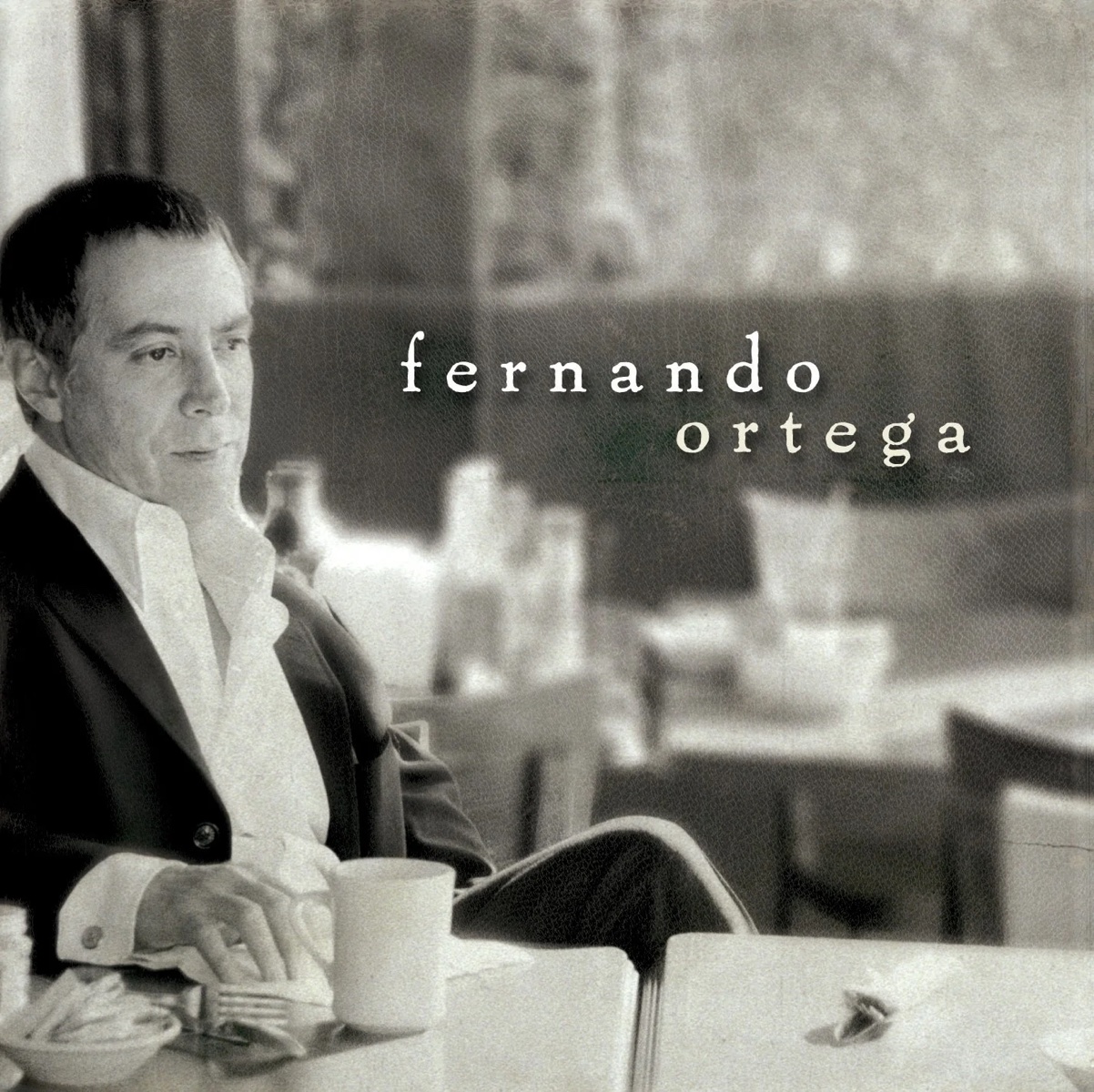 Album Art of Fernando Ortega's self-titled album - A black-and-white photo of a middle-aged man sitting at a table at a cafe or restaurant looking off into the distance with a coffee cup in front of him. Behind him are more empty tables and chairs. Over the background and to the right of his face is his name in white, lowercase letters with a shadow.