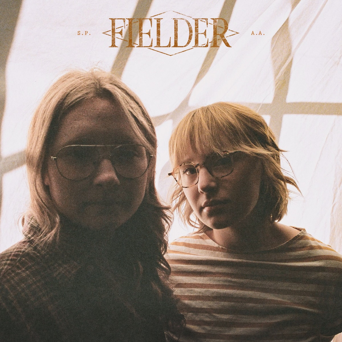 Album Art of Fielder's self-titled album - Lit mostly from behind through a curtain, two young women with blonde-brown hair and wearing brown colored tops are photographed from the chest up. The left one has shoulder-length hair, while the right one has a bit shorter. They both are wearing glasses and are looking towards the camera. Above their heads, printed in a golden brown color is a diamond design and the band name in a large, serif font.