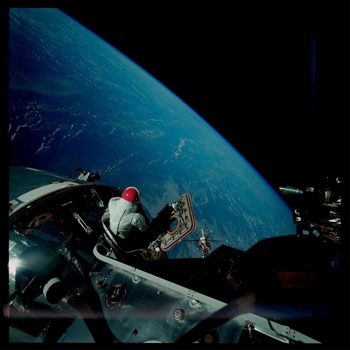 Album Art of Future Of Forestry's Sojourner album - A photo that looks to be taken from a space ship or space station orbiting Earth. The blue curve of Earth fills much of the background and in the foreground is the space station/ship. In one spot near the middle a person in a space suit is sitting inside a hatch with a red helmet, looking away from the camera towards Earth. There's a black border around the photo's edge.
