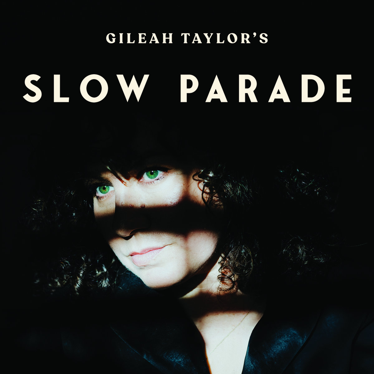 Album Art of Gileah Taylor's Slow Parade album - The top half is a black background with the words 'Gileah Taylor's' in small beige text centered near the top, then 'Slow Parade' in bigger text below it. On the bottom half, a photo of a a white woman with bright green eyes from the shoulders up on a dark background, with the top of her head, the middle of her face (the cheeks and the nose) as well as her neck are in bands of dark shadow.