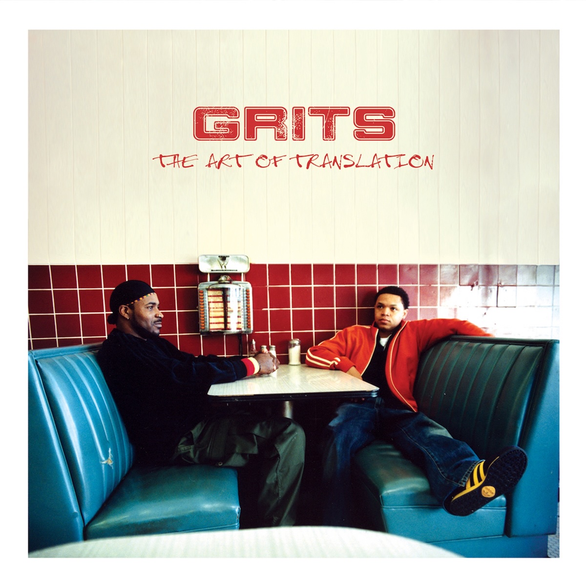 Album Art of GRITS's The Art of Translation album - With a thick white border, a photo of two black men sitting in blue padded benches of what looks like a very standard American diner. The wall behind them is dark red tile, and the table is white. There is a Nickelodeon, a machine to request songs from the jukebox, on the wall next to the table, and a few condiments as well on the table. The two people are sitting on opposite sides of the table and are not looking towards the camera. On the beige wall era above the tile, the group name is printed in red in a blocky text. And below it in a script font is the album name.