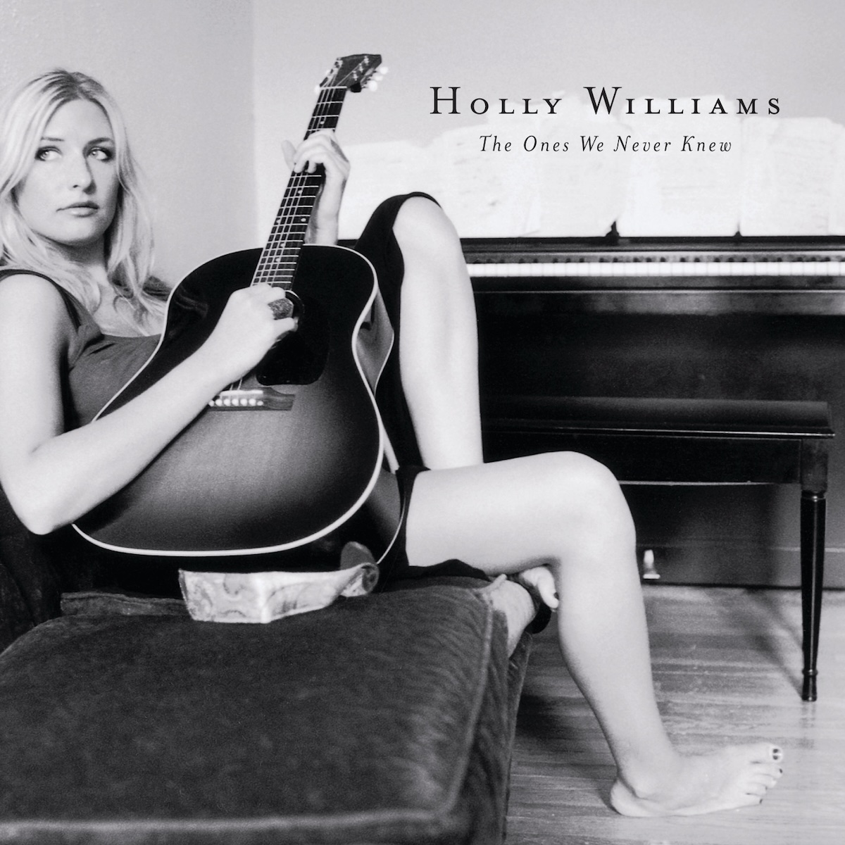 Album Art of Holly Williams's The Ones We Never Knew album - A black-and-white photo of a woman slouched on a couch with a large guitar on her lap, strumming it and looking to the left of the camera. The couch and her is mostly on the left side of the frame, with her right leg extending to the floor to the middle of the frame. The foot is on the wooden floor and behind her can be seen an upright piano with music spread out over the top of it. Where the piano meets the wall, printed above the photo is the artist's name and in smaller print, the album title below that.