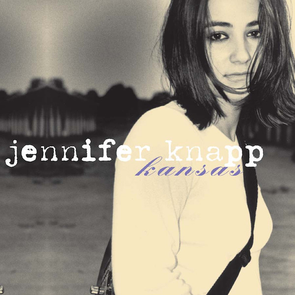 Album Art of Jennifer Knapp's Kansas album - A black and white photo of a woman standing it what could be a field with trees in the background, though it's hard to make out. The woman has dark shoulder-length hair and is wearing a light-colored shirt. She has a guitar on her back and the strap crossing her front chest. She is looking into the camera with a somewhat blank expression.