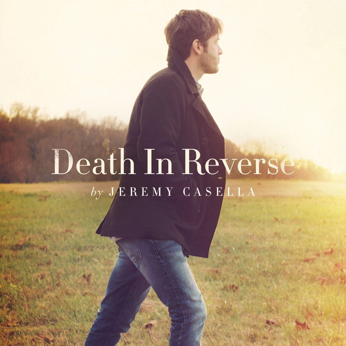 Album Art of Jeremy Casella's Death In Reverse album - A photo of a white man striding across a grassy field with a line of trees off in the distance. The man is striding to the right and is wearing blue jeans and a black coat, and has brown hair. he is looking away from the camera towards the sunset, which seems to be just around the right edge of the frame near the horizon. On top of the photo in the center is the album title in large, creamy white text, with 'by Jeremy Casella
