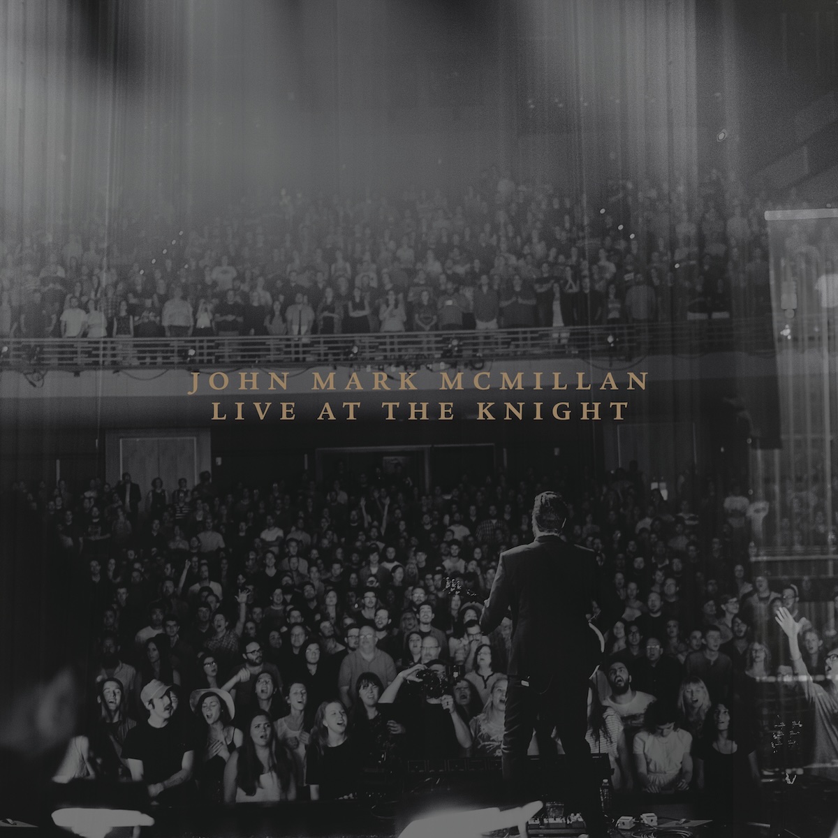 Album Art of John Mark McMillan's Live At The Knight album - A black-and-white photo of a man on a stage in the foreground, facing away from the camera and towards an audience. He's wearing a suit coat and holding a guitar. The audience is on the floor and on a balcony, hundreds and hundreds of people. Obscuring the video on the top and on the right is some sort of light refracting in the lens or some sort of effect to make it look cooler, mostly vertical lines of light. In the middle of the photo is printed in a brownish-gold serif font the album name on one line and then the artist name on a line below.