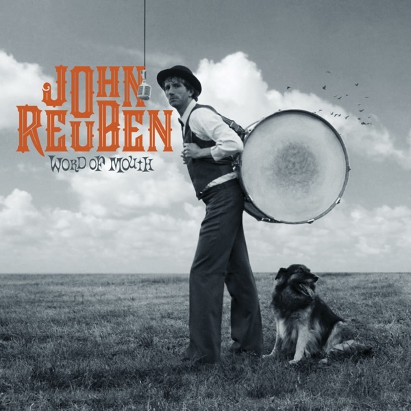 Album Art of John Reuben's Word Of Mouth album - A black and white photo of a man with a large kick drum slung over his shoulder on to his back is in a field and looking to the left. A microphone is hanging from the top of the frame to the left of him. A dog is sitting behind him and looking to the right, away from the man. To the left of the man on the top left is the artist's name in large orange type, and the album title in much smaller text in gray type.