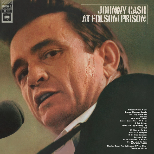 Album Art of Johnny Cash's At Folsom Prison album - A photo of Johnny Cash's face from below, with a microphone in front of his mouth. You can see he's wearing a black suit coat and a white shirt underneath, and that his guitar strap is going over his shoulder. He's looking right at the camera with an expression that may be him in the middle of talking. Beads of sweat can be seen on his cheek. In the top right the artist and album name is printed in a block font.