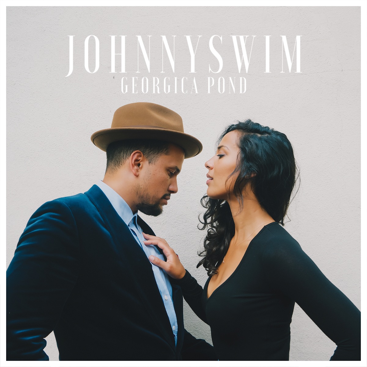 Album Art of Johnnyswim's Georgica Pond album - In front of a beige-white painted wall, two people are facing each other and very close together. The man is on the left and is looking down to the woman's chest, while the woman is looking up at his forehead. He's wearing a brown brimmed hat and has a navy blue suit and a powder blue shirt on with no tie. She is wearing a black top with sleeves and has he right hand on his chest. She is black but fair-skinned and he looks to be Latino with a short beard. Above them is printed in thin, tall white text the duo's name in large letters, with the album title in smaller letters. There's a white border around the edges.