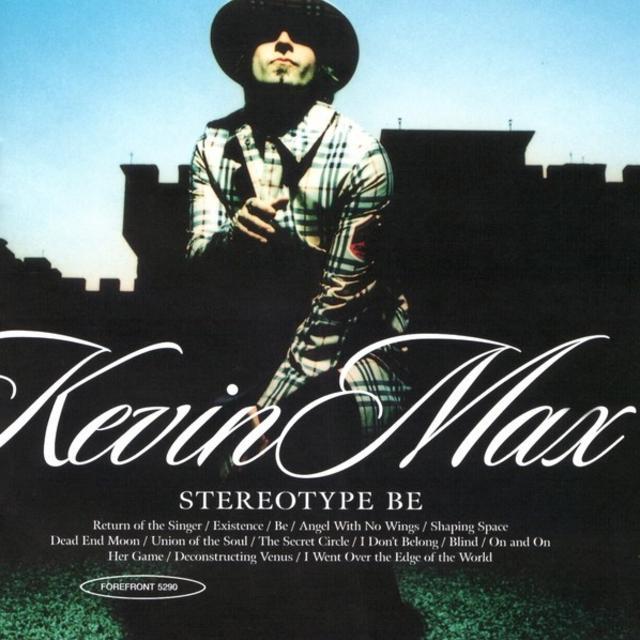 Album Art of Kevin Max's Stereotype Be album - A photo of a man looking at the camera and lit from the front, with a shadow of a building and a blue sky behind them. A bit of the green grass can be seen from the lighting as well, but much of the middle background is in the shadow of the building. The man is wearing a large-brimmed hat that is not lit, and it shades his eyes and forehead from view. We can see his nose, lips and goatee, They are wearing a stylish striped coat of mostly a beige color with black and red stripes. His right hand is reaching towards the camera. The artist name is in large, decorative letters in white. Below that is the album title in a smaller, simpler font. Below that in smaller letters is the track titles and the label catalog number in an oval printed over the photo in white.