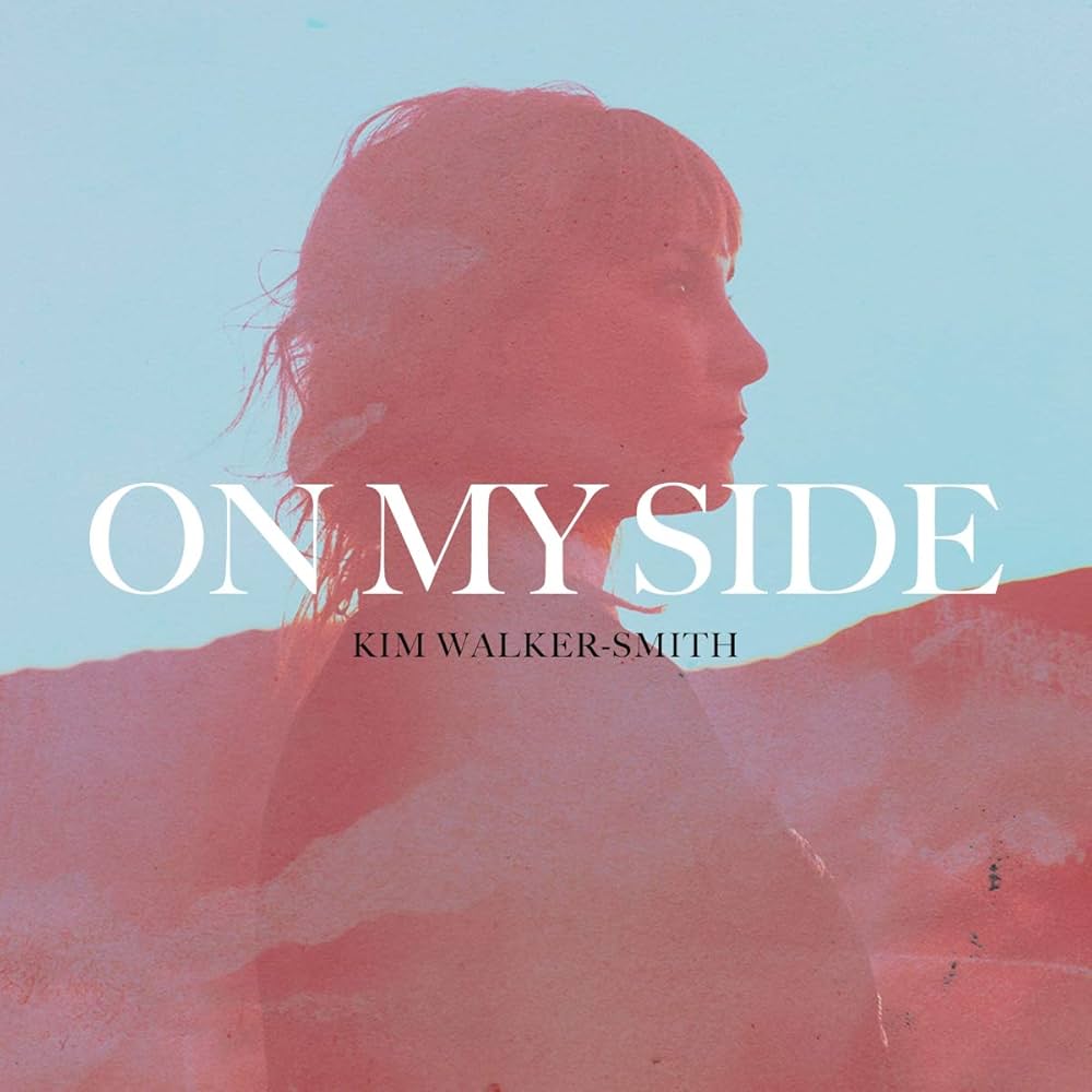 Album Art of Kim Walker-Smith's On My Side album - Over a heavily edited photo of a blue sky and a hillside and ground colored red, a profile shot of the artist's bust is superimposed. In large white letters is the album title, 'On My Side' and then in smaller black letters is the artist's name.