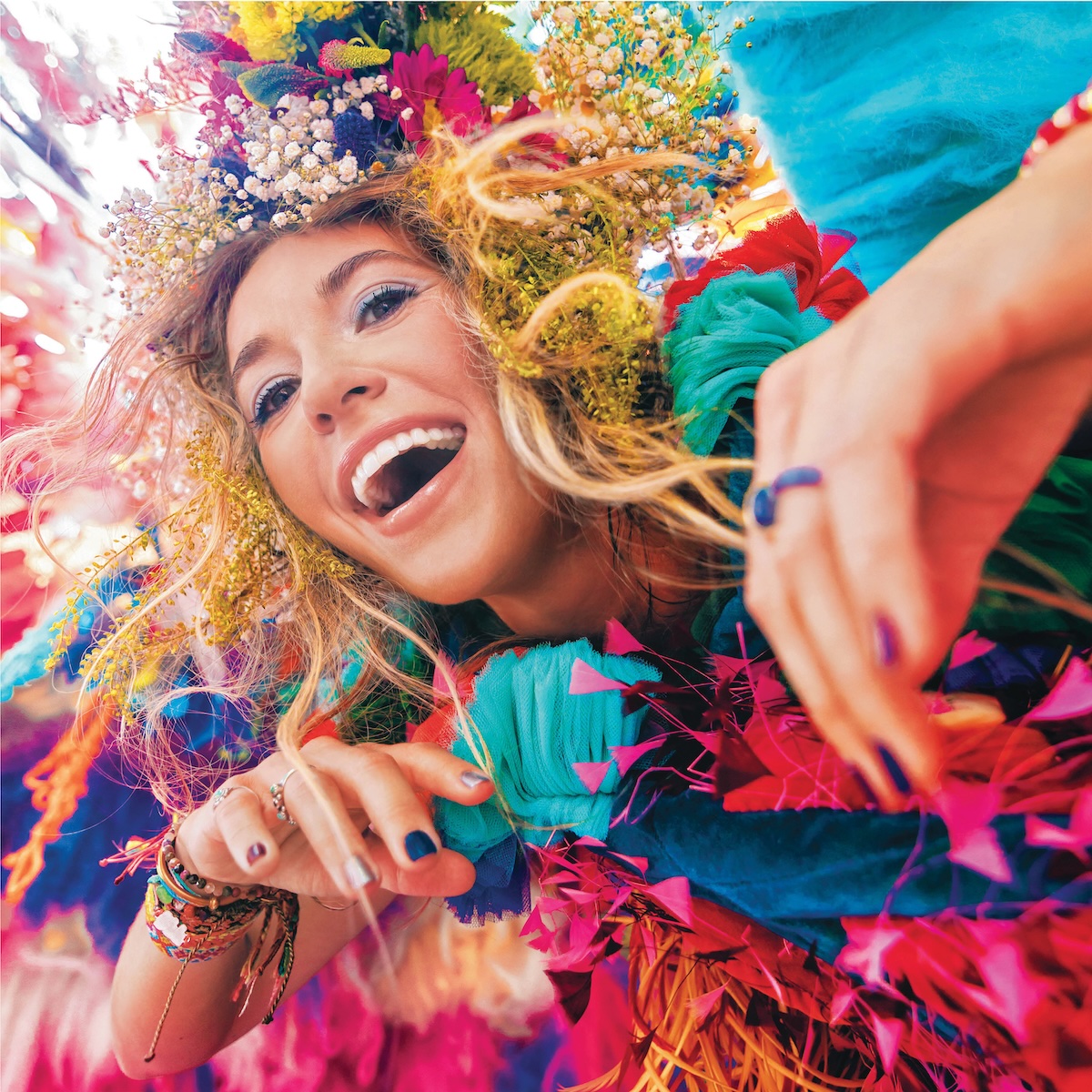 Album Art of Lauren Daigle's self-titled album - A photo of a young woman peering into a camera with a gaping wide smile. Her hands, each nail painted a different color, are near her chest and reaching towards the camera. On her head, mixed in with her brownish-blond hair is a bunch of flowers and feathers, it seems, of all colors. Whatever she seems to be wearing for clothes is a frilly, feathery, colorful mess and the background seems to be just as colorful and bright, like she may in the middle of a parade of many colors or something.