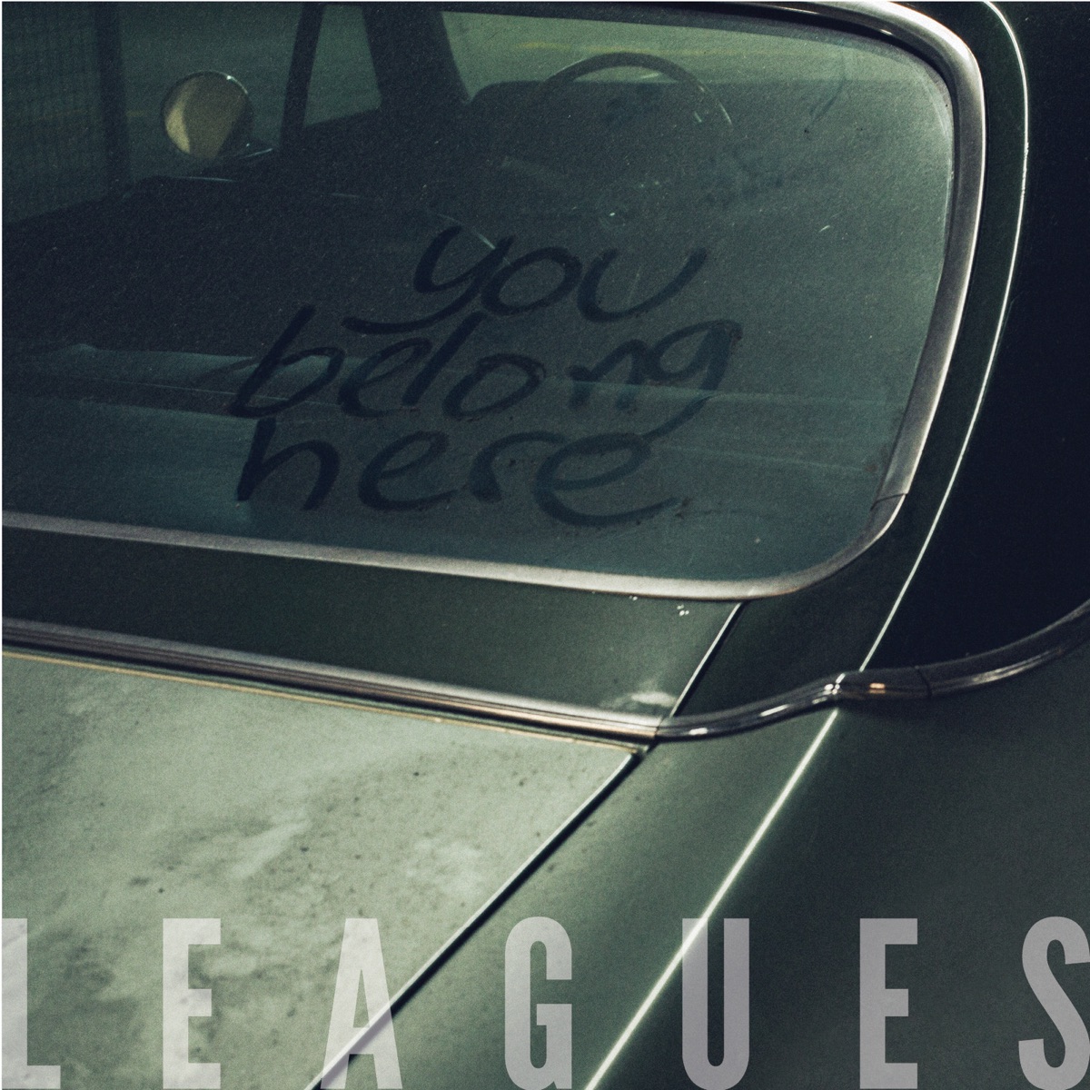 Album Art of Leagues's You Belong Here album - The back and side of a classic car of some sort, a green color with chrome edging. The car is very worn, with the paint coming off the top of the trunk. The window into the car is dusty and someone wrote 'you belong here' in the dust with their finger.