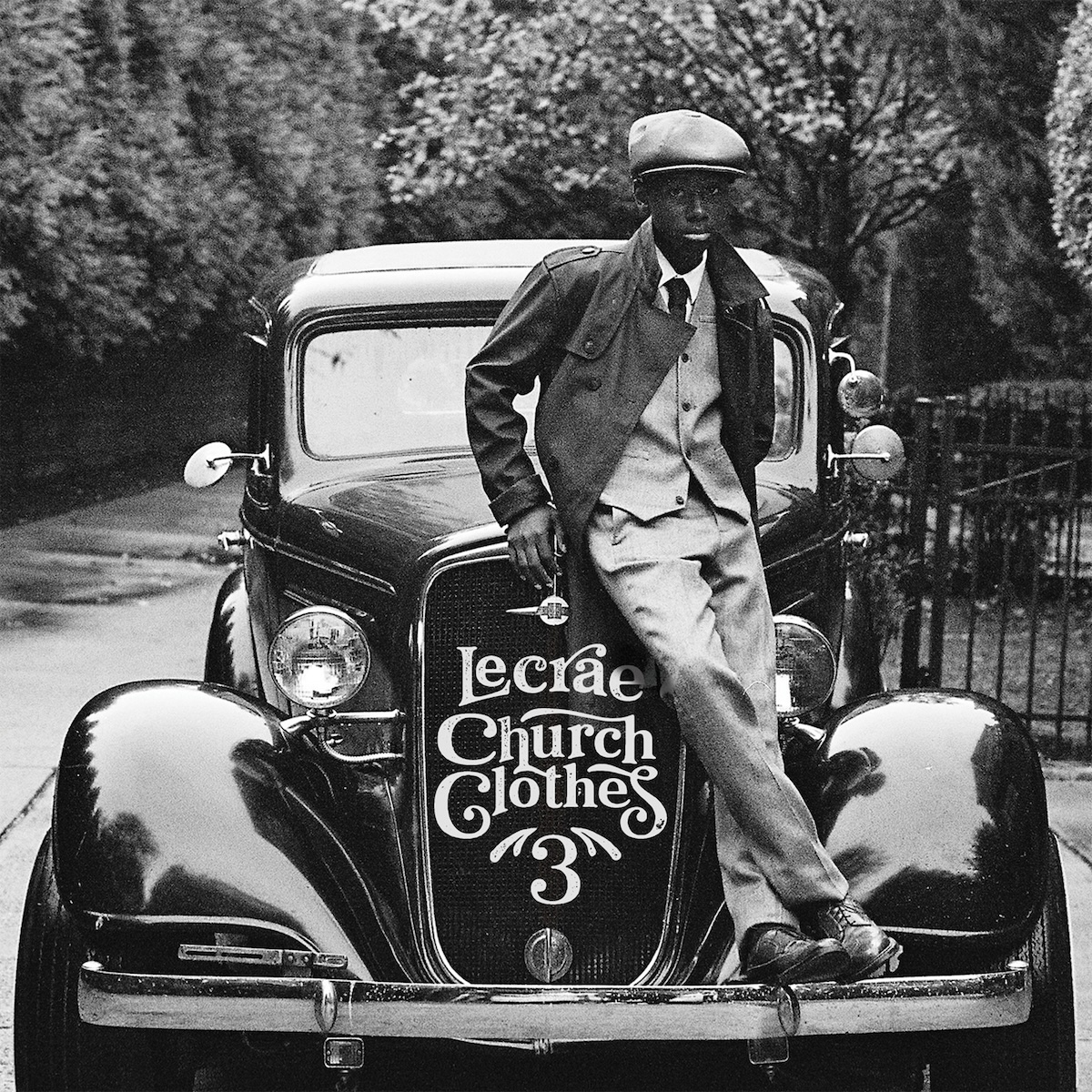 Album Art of Lecrae's Church Clothes 3 album - A black and white photo of a black kid standing on the front of a vintage, early 1900s car's bumper and sitting on the edge of the hood. He's also in a vintage coat with a vest and pants of a lighter shade and a white shirt and dark tie underneath. The car is sitting on cement near a wrought-iron fence and there's trees in the background. Where the large front grill is, printed in front of it is the artist name and album title in a somewhat decorative font in a light grey color.