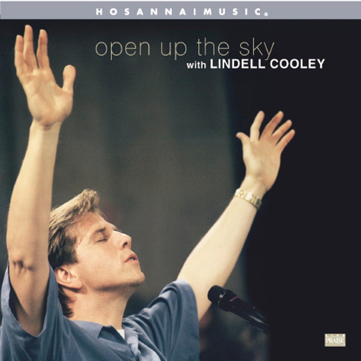 Album Art of Lindell Cooley's Open Up The Sky album - A photo of a middle-aged white man with his hands raised to up the heavens in a short-sleeved polo shirt is standing in front of a microphone. The background is dark black and gray, out of focus--probably a blank wall. Towards the top, it has the album title in grey and the line 'with Lindell Cooley' printed below it in smaller white letters. Across the top there is a powder blue bar with the words 'Hosanna Music' printed in the middle.