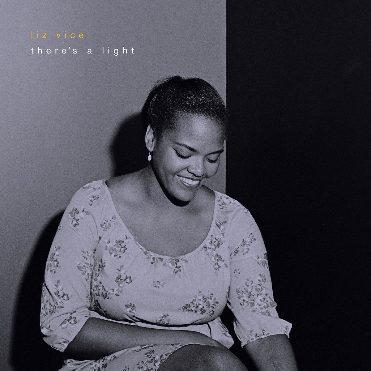 Album Art of Liz Vice's There's A Light album - A black-and-white photo of a woman sitting in a chair with a bright light hitting her from her front, off to the right and behind the camera. There's a grey background behind her, and off to the right it's just black, with a shadow of her body on the grey background. She's wearing a dress with a flower print and has has her hair pulled back behind her head. She is looking down towards her lap and has a big, toothy smile. In the top left, it has the artist's name in gold print in a small font, and below it the album name.