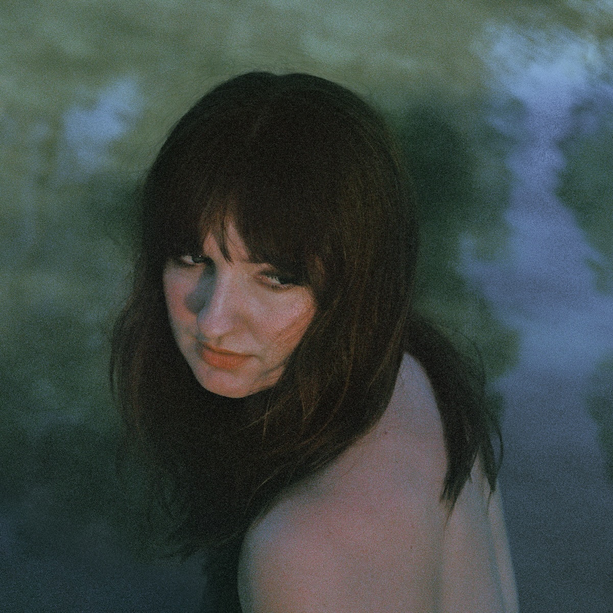 Album Art of Madison Cunningham's Ace album - A photo of a woman from slightly above in front of or in some sort of body of water, as the trees and sky are reflecting off the water behind her. The photo is pretty grainy, but her dark brown hair is covering her shoulders. She is facing away from the camera but looking back over her shoulder. We see her bare upper back and shoulder in the bottom of the frame. She is looking to the far right of the frame, in the direction of the sun, and has a blank expression on her face.