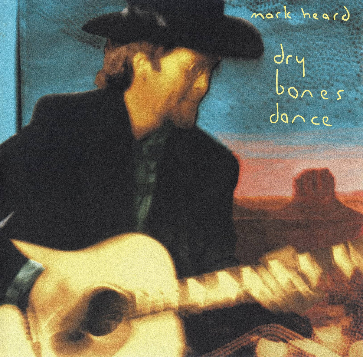Album Art of Mark Heard's Dry Bones Dance album - A man in a black cowboy hat and a black jacket is on the left side, playing a guitar. The man is in motion so the photo of him is a bit blurry, He has a brown beard and long brown hair and small circular, wire-rimmed glasses on his nose. Behind him can be seen a blue sky and a landscape of the western US, maybe Devil's Tower being visible off to the right over the top of his guitar neck. Handwriten in the top corner is the artist name in smaller yellow letters and the album title in slightly bigger letters.
